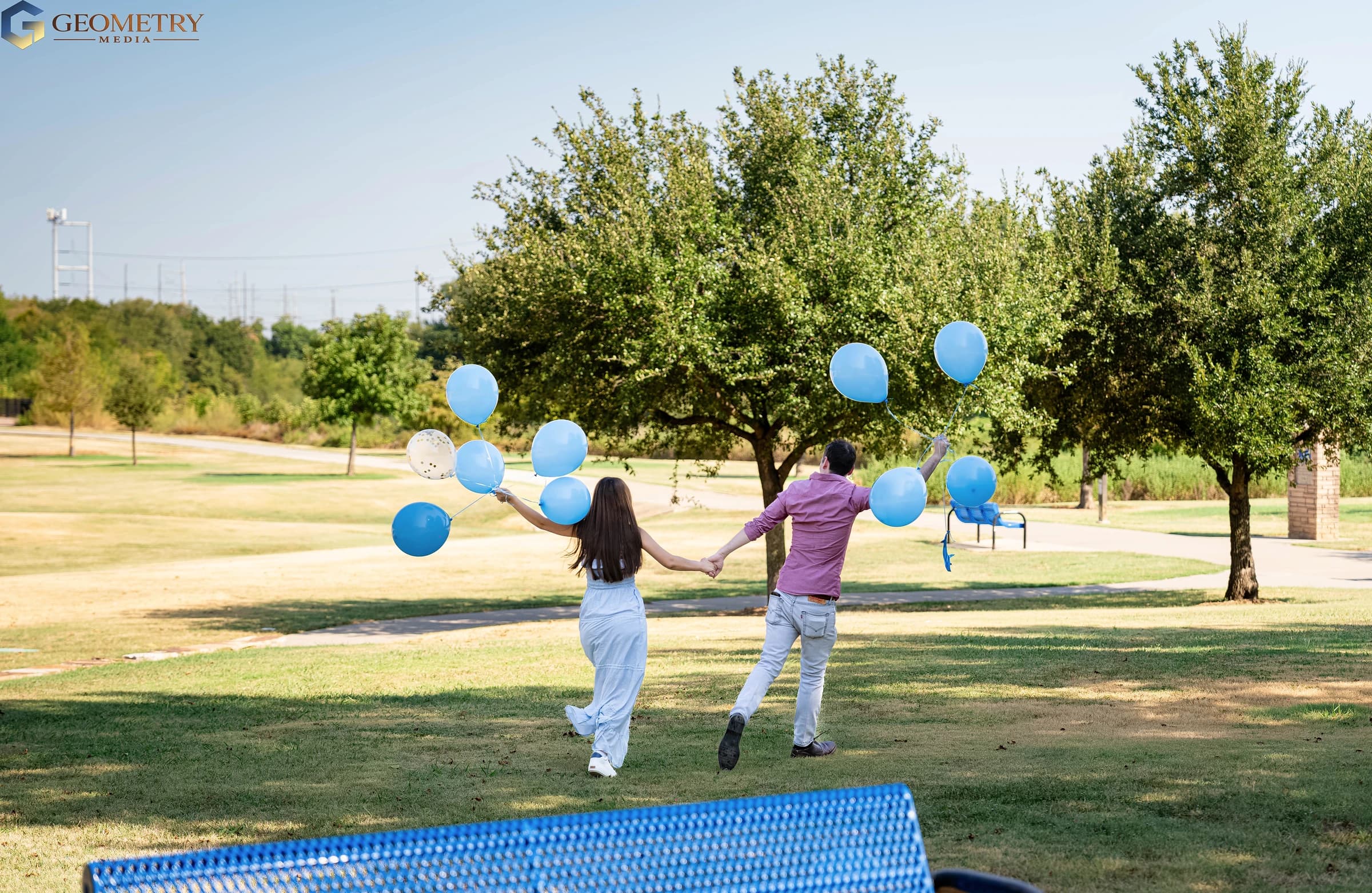 Hand in hand, the couple walk beneath the trees with blue balloons